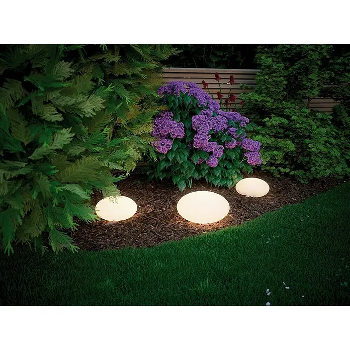 Paulmann Plug & Shine LED-Dekoleuchte Stone - 2,8 W, Weiß, L x B x H: 35 x 35 x 19,4 cm - Stimmungsvolle Gartenbeleuchtung: Drei runde Gartenleuchten erhellen ein Blumenbeet mit lila Blumen und Sträuchern.