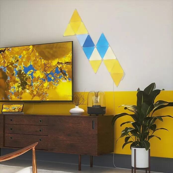 Nanoleaf Starterkit Shapes Triangles 15 Panels - 15 Stk., Weiß, RGBW, Triangles, Smart Home-fähig: Ja - Modulare, geometrische LED-Wandleuchten in Gelb und Blau, dynamisch über einem Fernseher und einer Anrichte in einem modernen Wohnzimmer angeordnet.