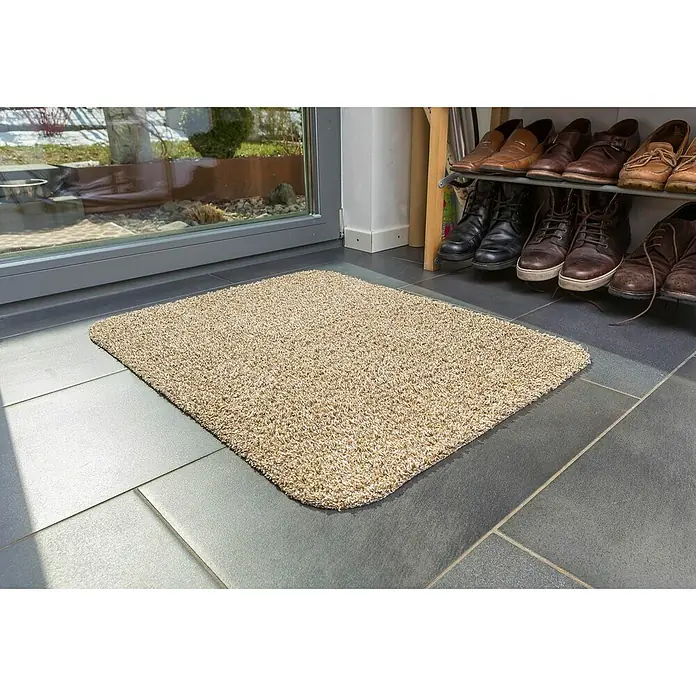 Astra Fußmatte Entra Saugaktiv - Beige, 75 x 130 cm, Material Nutzschicht: 100 % Baumwolle - Ein beiger, rechteckiger Teppich liegt vor einer Tür, neben einem Schuhregal mit verschiedenen Schuhen darauf. Der Teppich sorgt für Sauberkeit und Komfort.