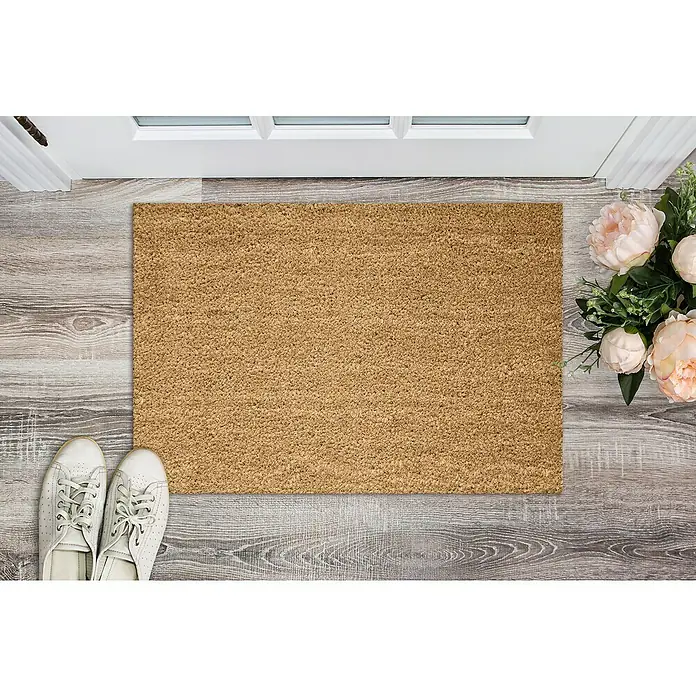 Astra Fußmatte - 40 x 60 cm, Natur, Material Nutzschicht: 100 % Kokos, Höhe: 24 mm - Eine rechteckige, hellbraune Fußmatte liegt vor einer weißen Tür, neben weißen Schuhen und rosa Blumen. Sie dient als dekoratives Element.