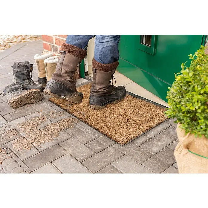 Astra Fußmatte - 50 x 80 cm, Natur, Kokosvelours, Kokos - Fußabstreifer vor einer grünen Tür mit schmutzigen Stiefeln darauf, flankiert von einem Paar Stiefel und einem Strauch.