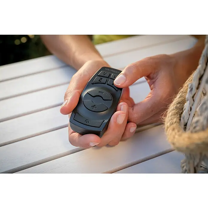 Paulmann Plug & Shine Controller mit Fernbedienung - Max. Leistung: 75 W, Schwarz, IP68 - Nahaufnahme einer Person, die eine schwarze Fernbedienung hält, die auf einem weißen Tisch liegt und eine einfache Steuerung ermöglicht.
