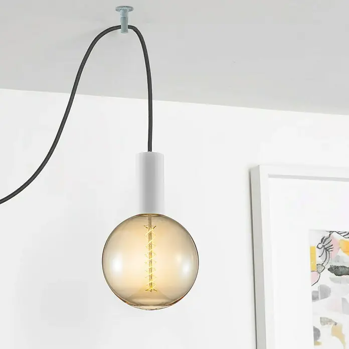 Home Sweet Home Deckenhaken - Beton, Ø x H: 3,5 x 5 cm - Nahaufnahme einer hängenden Lampe mit Glühbirne, die ein warmes Licht ausstrahlt, an einer weißen Wand mit einem Kunstwerk im Hintergrund.