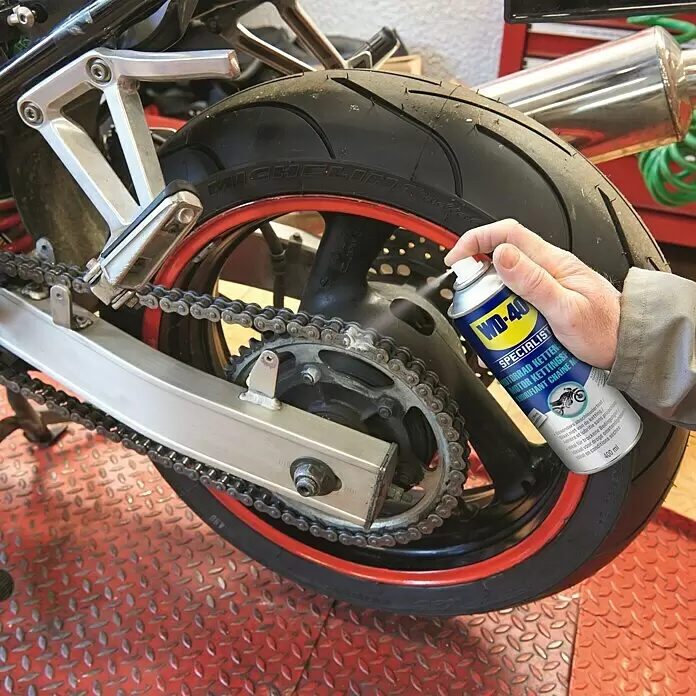 WD-40 Specialist Motorbike Kettenspray Classic - 400 ml - Eine Hand sprüht blau-gelbes WD-40 Spezial-Kettenspray auf die Kette und das Hinterrad eines Motorrads.