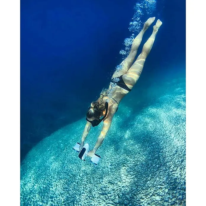 Sublue Tauchscooter White Shark - L x B x H: 465 x 230 x 230 mm - Eine Frau taucht mit Unterwasser-Hanteln im blauen Meer. Die Hanteln sind weiß mit schwarzen Akzenten und sie scheint ein Unterwasser-Workout zu machen.