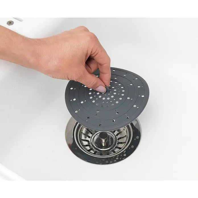 Wenko Haarfangsieb - Durchmesser: 12 cm, Silikon, Grau - Eine Hand setzt einen grauen Spülen-Sieb über einen glänzenden Edelstahl-Abfluss in einem weißen Waschbecken ein.