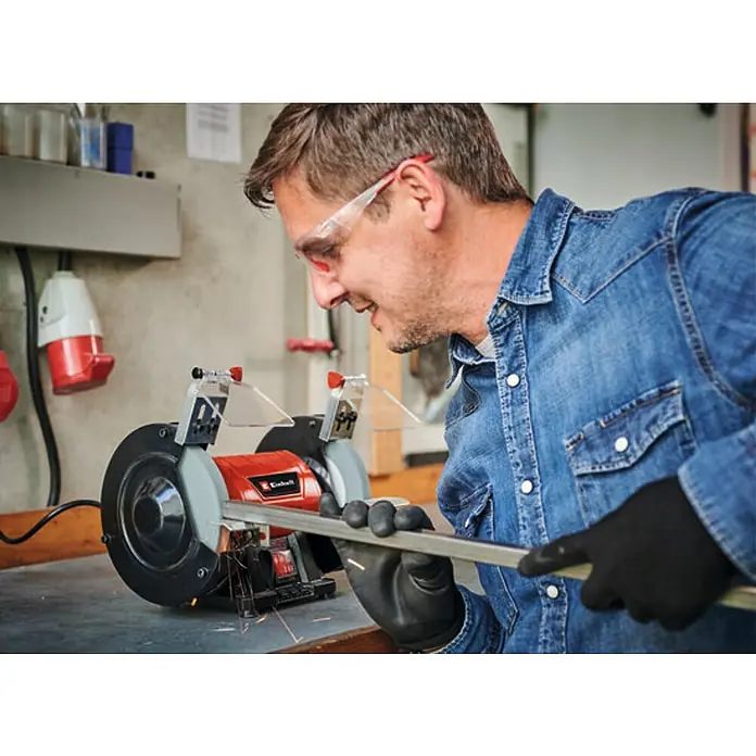 Einhell Esmeriladora doble eléctrica TC-BG 150 - 150 W, 2.980 r.p.m. - Un hombre usa una amoladora de banco Einhell para trabajar una pieza de metal, usando gafas de seguridad y guantes para protegerse.