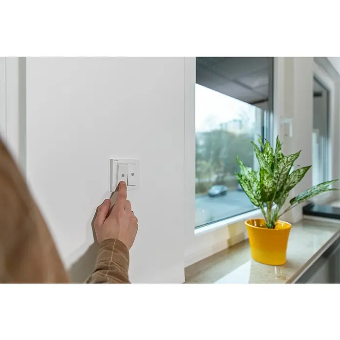 Schellenberg Interruptor para persianas - Interruptor basculante doble, Específico para: Persianas, toldos y estores motorizados - Una mano presiona un interruptor de luz blanco moderno en una pared blanca, junto a una ventana con una planta en maceta.