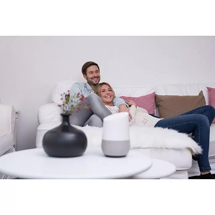 Schellenberg Recogedor de cinta de persiana inhalámbrico Rollodrive 65 Premium - Fuerza de tracción: 45 kg, Anchura de la correa: 23 mm, Empotrado - Un altavoz inteligente blanco y gris en una mesa de centro, integrado en una cómoda escena de salón con una pareja relajándose.