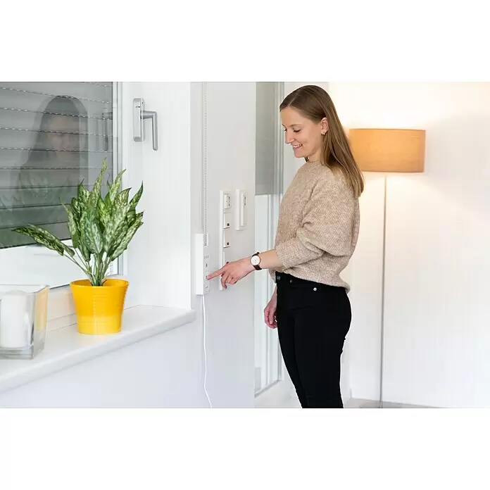 Schellenberg Recogedor de cinta de persiana inhalámbrico Rollodrive 65 Premium - Fuerza de tracción: 45 kg, Anchura de la correa: 23 mm, Empotrado - Una mujer presiona un botón en un panel de control domótico blanco montado en la pared junto a una ventana.
