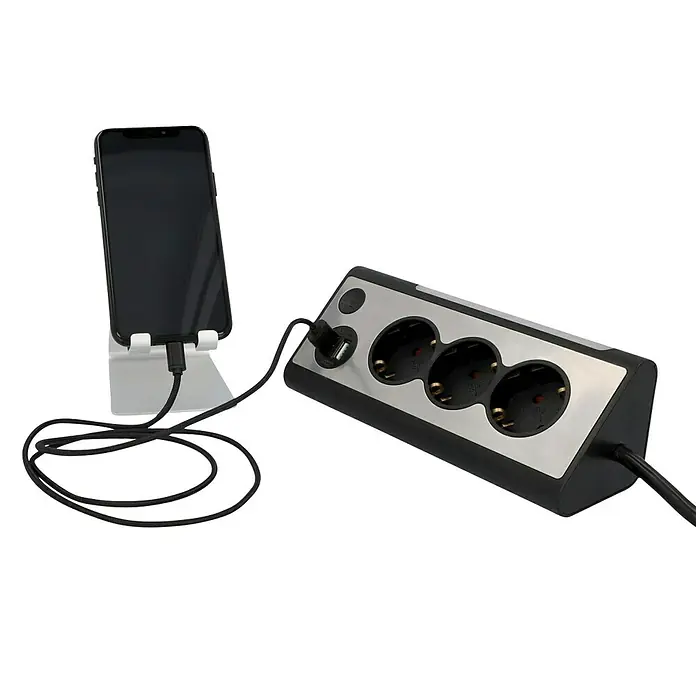 Tischsteckdose - 3-fach, Silber/Schwarz, 28 x 10,2 x 9 cm - Eine Steckdosenleiste mit drei Steckplätzen und einem USB-Anschluss, an dem ein Smartphone zum Aufladen angeschlossen ist.