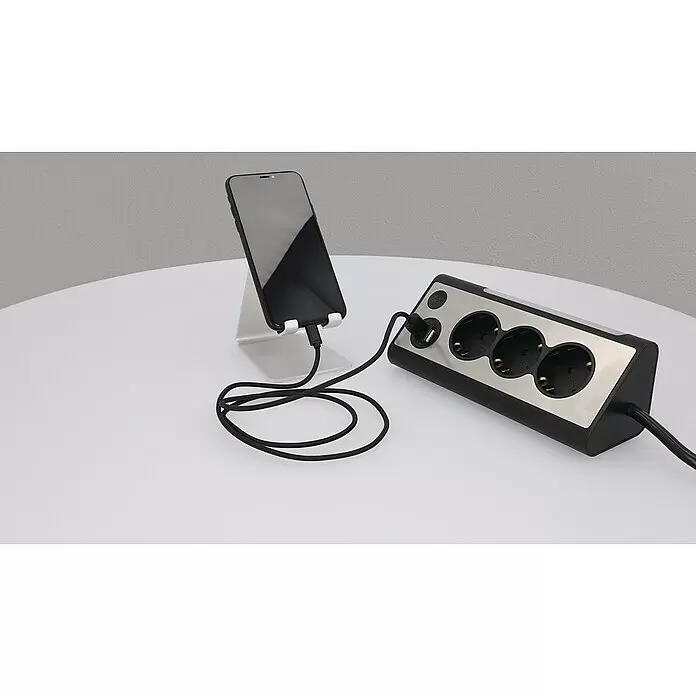 Tischsteckdose - 3-fach, Silber/Schwarz, 28 x 10,2 x 9 cm - Schwarze und silberne Steckdosenleiste mit drei Steckdosen und einem USB-Anschluss lädt ein schwarzes Smartphone auf einem silbernen Ständer.