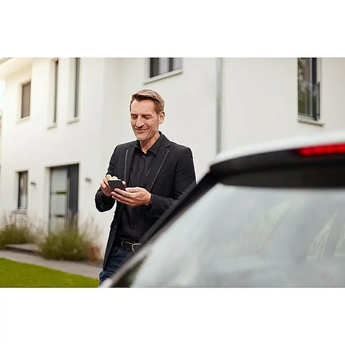 Homematic IP Alarmsirene HmIP-ASIR-2 - Batteriebetrieben, Alarmsignal: 90 dB, Ø x H: 12,4 x 4,5 cm - Ein stilvoller Mann in einem schwarzen Blazer und Jeans bedient sein Smartphone neben einem Auto, vor einem modernen Wohnhaus.