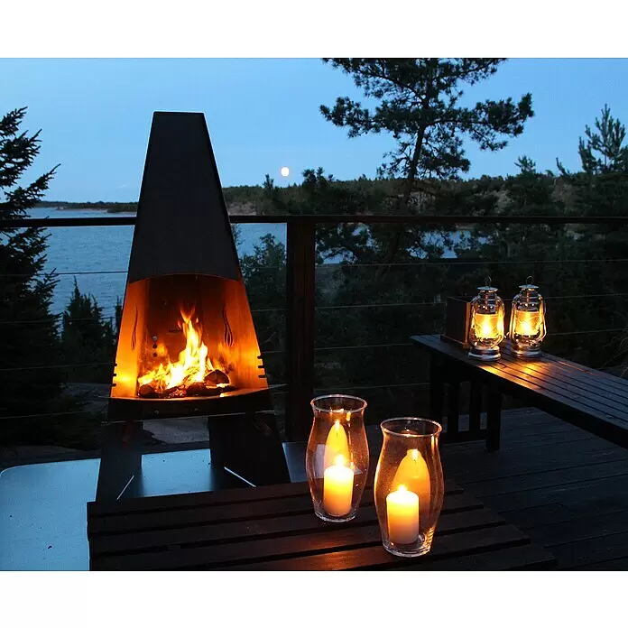 Aduro Gartenkamin - 58 x 60 x 124 cm, Stahl - Ein Outdoor-Metallkamin mit warmem Feuer, zwei Sturmlampen und zwei Kerzenhaltern aus Glas beleuchten eine Terrasse mit Seeblick bei Dämmerung.