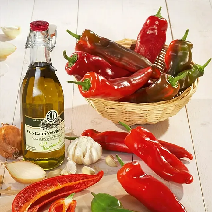 Piardino Peperoni 'Amboy' - Capsicum annuum, 12 cm, Erntezeit: Juli - Eine Flasche Olivenöl extra vergine, Paprika, Zwiebeln und Knoblauch liegen auf einem Tisch.