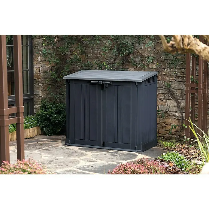 Keter Truhe Store it out Prime - L x B x H: 132 x 71,5 x 113,5 cm, Grau/Schwarz, Kunststoff - Außenaufnahme eines dunkelgrauen Keter-Gartenschranks mit Doppeltür vor einer Steinmauer. Praktische Gartengeräteaufbewahrung.