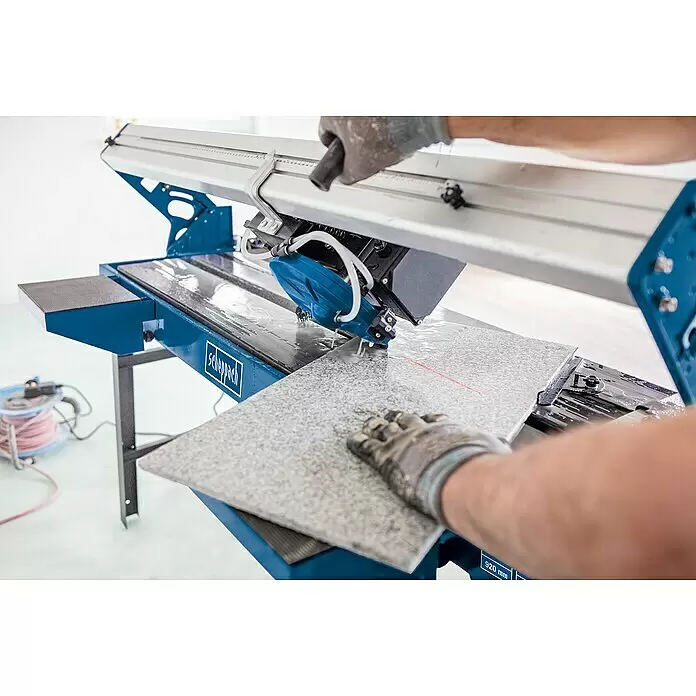 Scheppach Elektro-Fliesenschneider FS3600 - 900 W, Max. Schnittlänge: 920 mm, Schnitttiefe bei 90 °: 36 mm - Eine Person schneidet eine graue Fliese mit einer blau-silbernen Fliesenschneidemaschine, Wasser spritzt beim Schnitt.