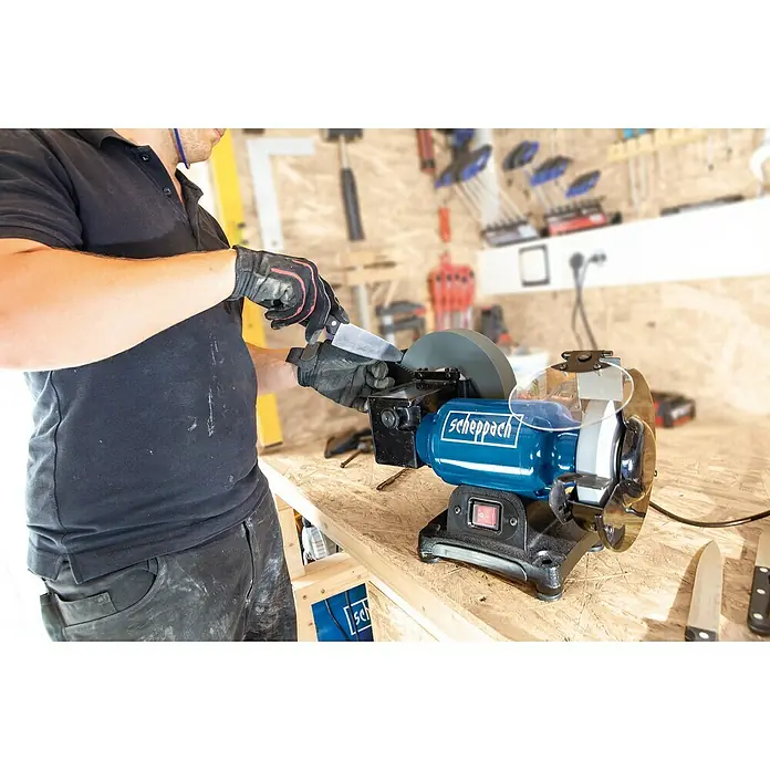 Scheppach Elektro-Nass-Trockenschleifer BG 200W - 250 W - Eine Person schärft ein Messer mit einer Scheppach Schleifmaschine auf einer Werkbank, umgeben von Werkzeugen.