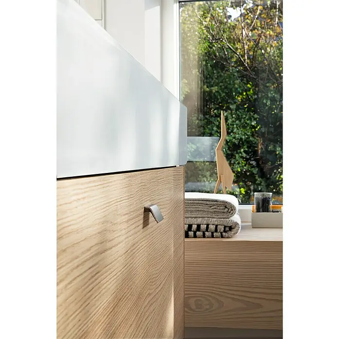 Duravit Vero Air Waschtisch - 100 x 47 cm, Ohne Spezialglasur, Hahnloch: Ohne, Ohne Überlauf, Weiß, Geschliffen - Nahaufnahme eines Badezimmerschranks mit Holzfront und modernem Griff, dazu Handtücher und Dekoration vor einem hellen Fenster.