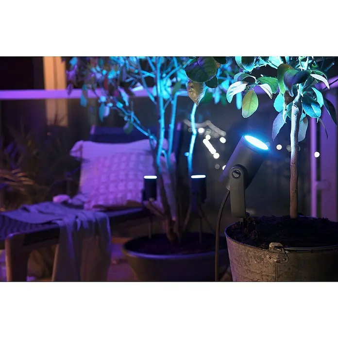 Philips Hue LED-Gartenspot Lily - 8 W, Lichtfarbe: RGB, IP65 - Nahaufnahme einer grauen Hue-Gartenleuchte, die einen Baum mit blauem Licht beleuchtet, und anderer Lichter im Hintergrund.
