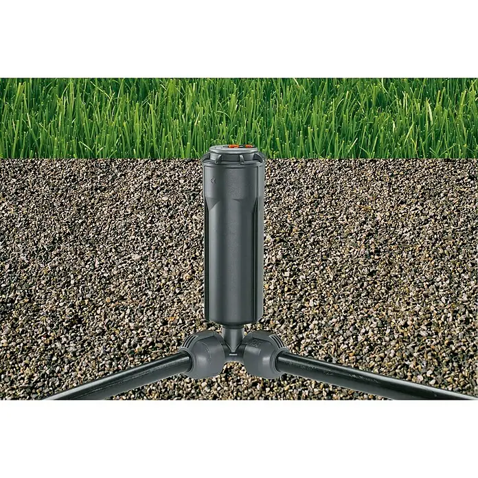 Gardena Sprinklersystem Codo angular - Diámetro: 25 mm, Dimensión conexión: ½″ - La imagen muestra un aspersor de césped retráctil instalado en un césped con piedras debajo.