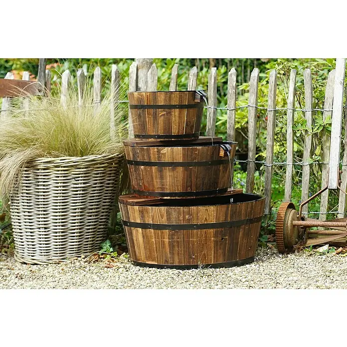 Ubbink AcquaArte Gartenbrunnen Edinburgh - Holz, Braun - Ein dreistufiger Holzfassbrunnen steht im Garten, neben einem Weidenkorb und einem alten Rasenmäher, der eine rustikale Atmosphäre schafft.