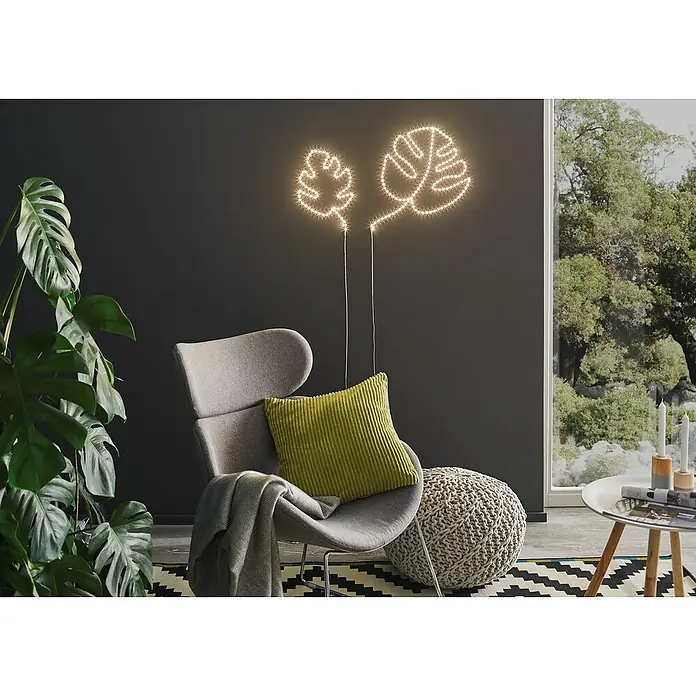 Paulmann LED-Band Function FlexLED - Länge: 5 m, Lichtfarbe: Warmweiß, 10,5 W - Elegantes Interieur mit Monstera-Blatt-Lichtern an einer dunklen Wand, ergänzt durch einen grauen Sessel und einen Strickhocker für gemütliches Ambiente.