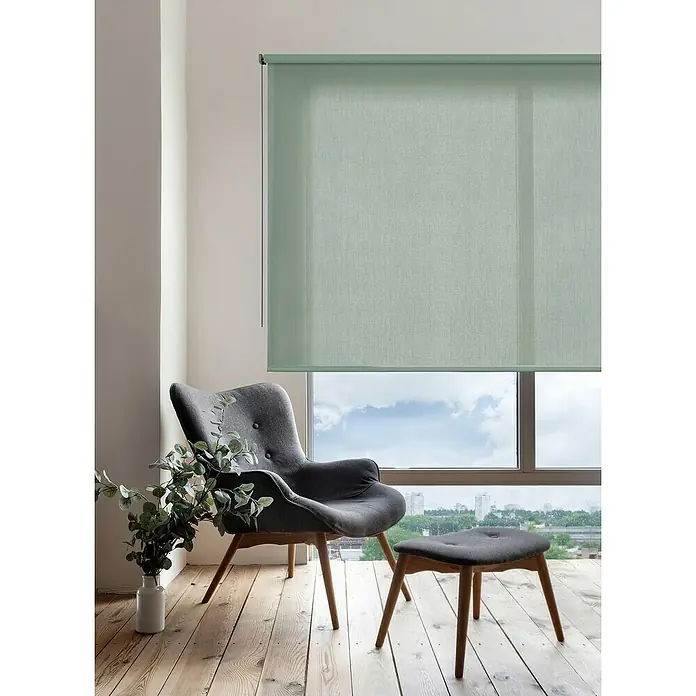 Estor enrollable Soft - An x Al: 80 x 250 cm, Eucalipto - Sillón gris y reposapiés junto a una ventana con persianas enrollables verdes, que ofrecen privacidad y luz suave.