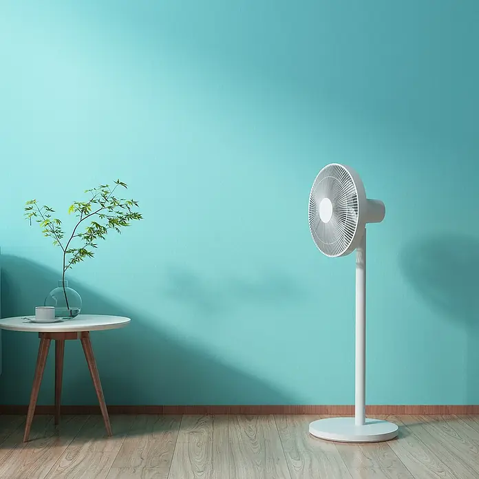 Xiaomi Ventilador de pie Smart Fan 2 - Blanco, 35,5 cm, 1.000 mm - Un ventilador de pie blanco sobre un piso de madera frente a una pared celeste, junto a una mesa con una planta y una taza.