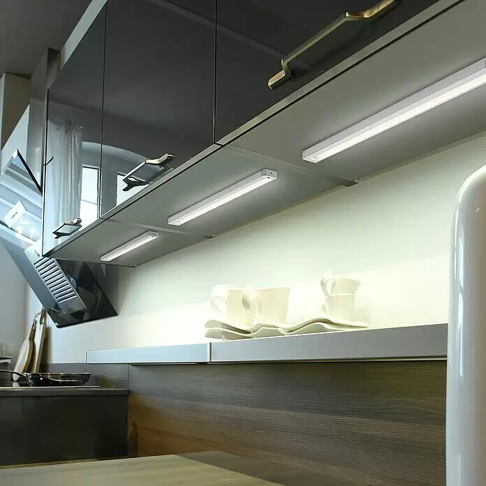 Paul Neuhaus LED-Unterbauleuchte - 6 W, Länge: 59,5 cm, Warmweiß - Weiße lineare LED-Unterschrankleuchten beleuchten eine moderne Küchenarbeitsplatte, hellen die Arbeitsfläche und gestapelte Tassen auf.