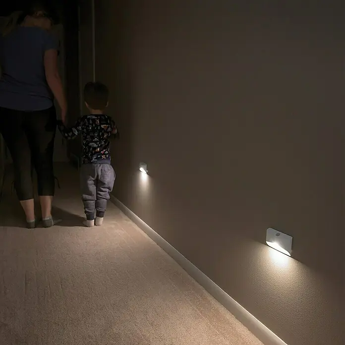 Mr. Beams LED-Nachtlicht MB710 - 15 lm, Lichtfarbe: Warmweiß - Wandmontierte LED-Leuchten beleuchten einen dunklen Flur, während eine Frau ein Kind hält und durch den Flur geht. Die Lampen sorgen für Sicherheit.
