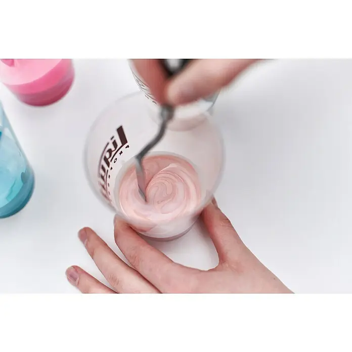 Liquitex Professional Gießmedium Irisierend - 473 ml, Transparent - Das Bild zeigt das Mischen von Farben in einem transparenten Behälter mit einem Rührwerkzeug, wobei die Farben rosa und weiß sind.
