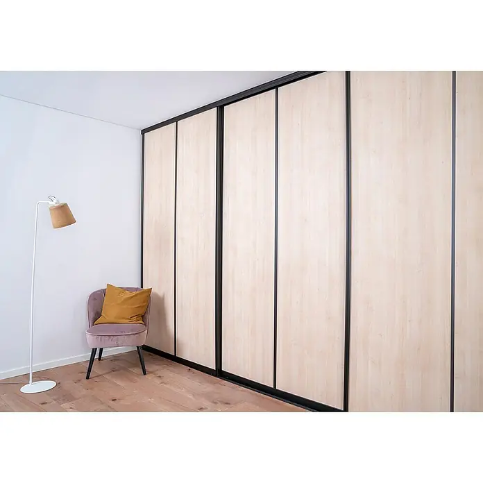 Room Plaza Easy Schiebetür-Bau-Set - Buche Naturwuchs/Ahorn Naturwuchs, Farbe Beschlag: Schwarz - Einbauschrank mit hellen Holzpaneelen und dunklen Rahmen in einem stilvollen Raum mit Sessel und Stehlampe.