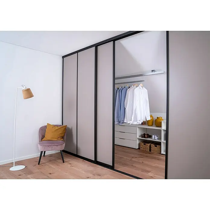 Room Plaza Easy Schiebetür-Bau-Set - Taupe/Weiß Miniperl, Farbe Beschlag: Schwarz - Elegantes Schlafzimmer-Interieur mit einem modernen Kleiderschrank mit Schiebetüren, einem stilvollen Sessel und einer Stehlampe.