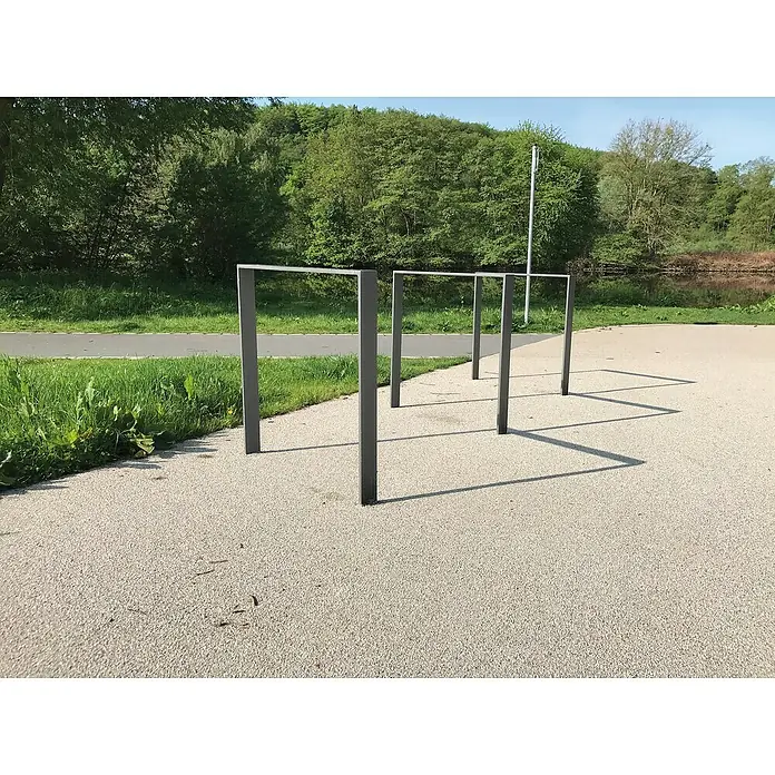 Schake Fahrrad-Anlehnbügel - B x H: 45 x 120 cm, Passend für: 2 Fahrräder, Beidseitige Radeinstellung - Eine Reihe von modernen Fahrradständern aus grauem Metall auf einem gepflasterten Platz vor einer grünen Landschaft. Sie werfen lange Schatten auf den Boden.