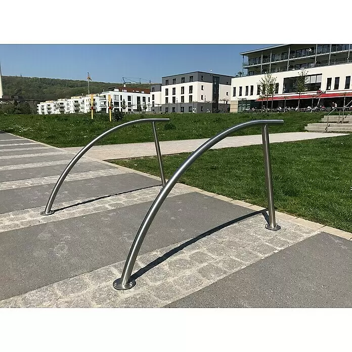 Schake Fahrrad-Anlehnbügel - B x H: 140 x 114 cm, Beidseitige Radeinstellung, Silber - Zwei silberne, geschwungene Metall-Fahrradständer auf einem gepflasterten Weg neben einer Grünfläche.