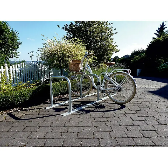 Schake Fahrrad-Anlehnbügel - B x H: 70 x 70 cm, Passend für: 6 Fahrräder, Beidseitige Radeinstellung - Ein weißes Fahrrad mit einem Korb am Lenker, geparkt an einem modernen Fahrradständer auf einem gepflasterten Weg.