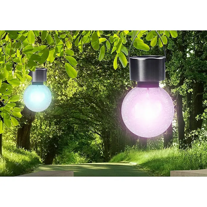 BAUHAUS Solar-Hängeleuchte - Nickel, Klar, IP44 - Zwei Solarleuchten mit Glaskugeln in Rosa und Blau hängen an Bäumen und erhellen einen grünen Gartenweg.