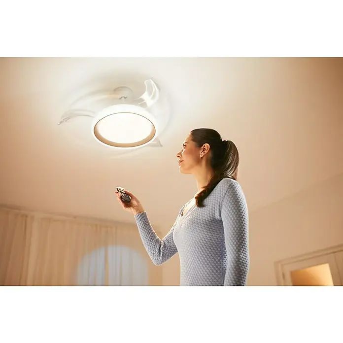 Philips Ventilador de techo Bliss Gold - Oro, 51 cm, 48 W, Blanco cálido - Mujer controla una moderna lámpara de techo con un mando a distancia, mostrando su iluminación brillante y uniforme.