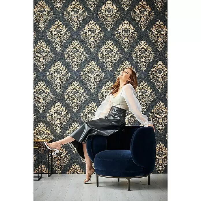 A.S. Création Metropolitan Stories II Vliestapete Damask - Blau/Gold, Ornament, 10,05 x 0,53 m - Dunkelblauer Samtsessel mit sitzender Frau, präsentiert luxuriösen Stoff und elegantes Design.