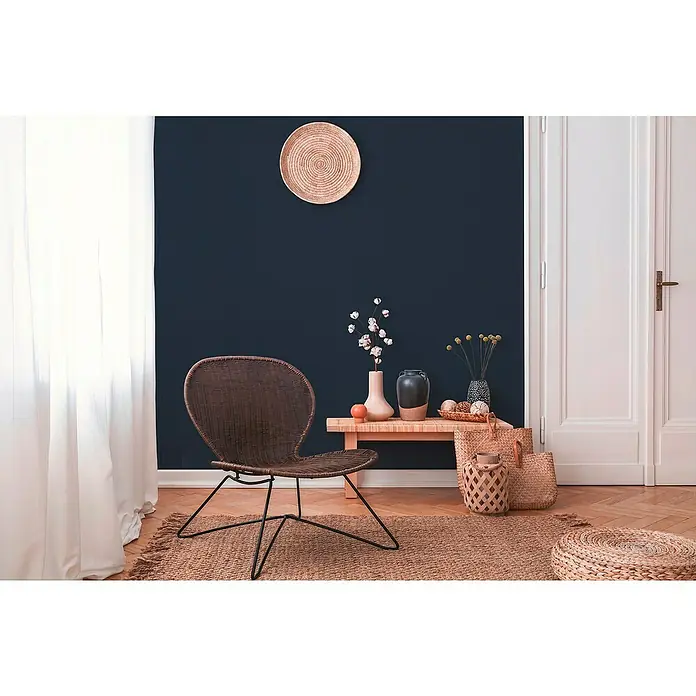 A.S. Création Metropolitan Stories II Vliestapete Textil-Uni - Schwarz, Uni, 10,05 x 0,53 m - Elegantes Wohnzimmer mit einem geflochtenen Stuhl im Bohème-Stil, der auf einem Teppich vor einer marineblauen Wand steht.