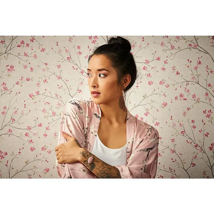 A.S. Création Metropolitan Stories II Vliestapete Kirschblüte - Creme, Floral, 10,05 x 0,53 m - Eine Frau mit Tätowierungen trägt einen rosa Kimono mit Blumenmuster und ein Armband mit rosafarbenem Stein.