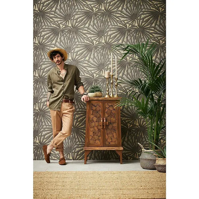 A.S. Création Metropolitan Stories II Vliestapete Palme - Grau/Gold, Floral, 10,05 x 0,53 m - Stilvolles Interieur mit einem Holzschrank, goldenen Kerzenständern und einer Palme, ergänzt durch einen lässig gekleideten Mann mit Hut.