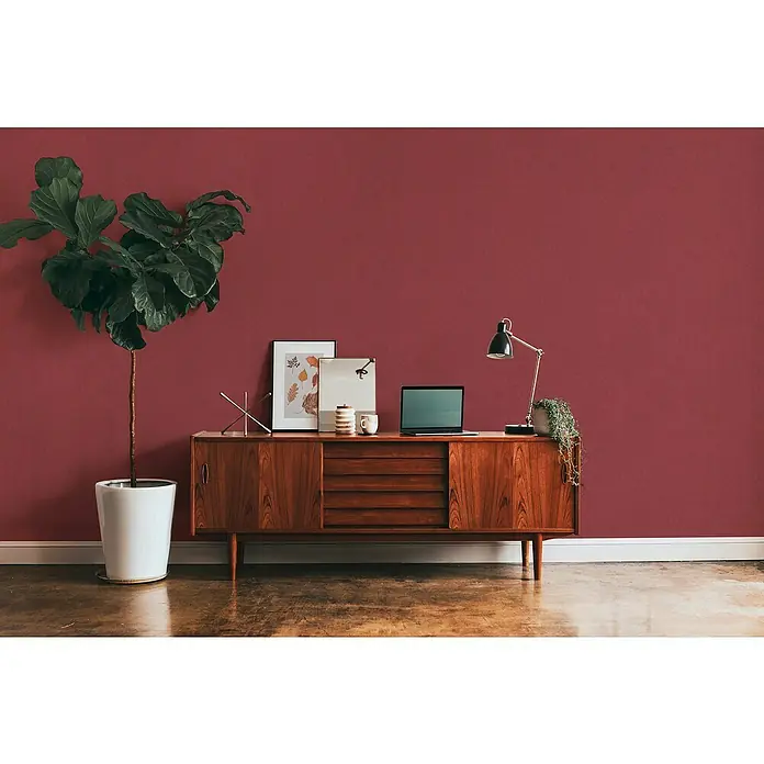 A.S. Création Metropolitan Stories II Vliestapete - Dunkelrot, Uni, 10,05 x 0,53 m, Matt - Elegantes Sideboard aus Holz mit dekorativen Elementen wie Lampe, Pflanze und Laptop, vor einer roten Wand.