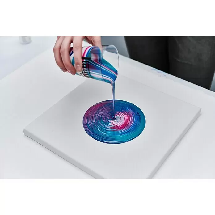 Liquitex Professional Gießmedium Matt - 473 ml, Transparent - Eine Hand gießt lebendige blaue, rosa und lila Acrylfarbe aus einem durchsichtigen Becher auf eine weiße Leinwand und erzeugt ein Wirbelmuster.