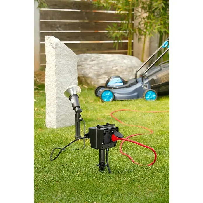 Voltomat Erdspieß-Steckdose - Kabellänge: 5 m, 4 Stk., IP44 - Nahaufnahme einer schwarzen Gartensteckdose mit rotem Stromkabel auf Rasen vor einem Garten.