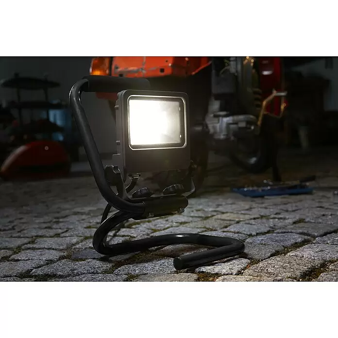 Ledvance LED-Strahler Floodlight - 20 W, 1.600 lm, 4.000 K, IP65 - Dunkelgraue LED-Arbeitsleuchte auf schwarzem Ständer, beleuchtet gepflasterten Garagenboden und Reparaturarbeiten am Roller.