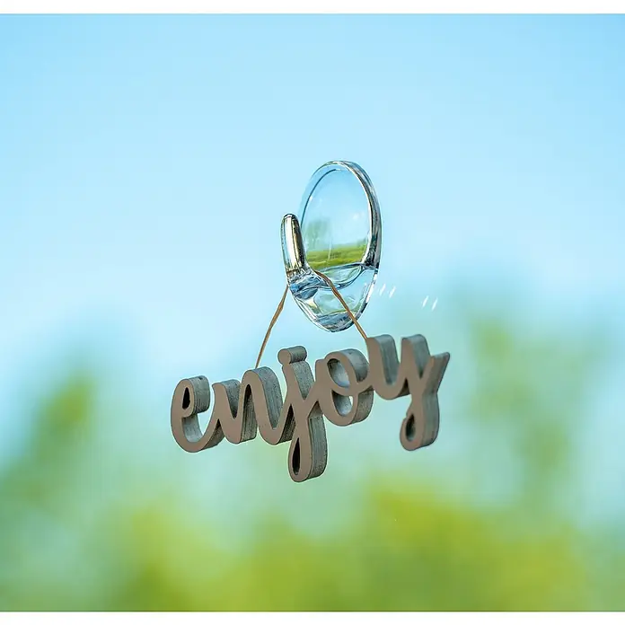 Wenko Handtuchhaken Groß - 2 Stk., Transparent, Kunststoff - Genießen: Nahaufnahme eines Schriftzugs "Enjoy", der mit einem transparenten Klebehaken an einer Scheibe befestigt ist, vor blau-grünem Hintergrund.