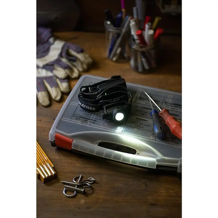 Ledlenser Stirnlampe H7R Core - 15 lm - 1.000 lm, IP67 - Eine Ledlenser Stirnlampe beleuchtet eine Werkzeugkiste auf einem Holztisch, umgeben von Werkzeugen und Handschuhen.