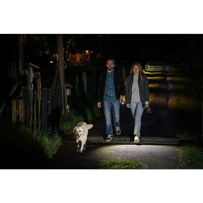 Ledlenser Taschenlampe P7R Core - 15 lm - 1.400 lm, IP68 - Ein Paar geht nachts mit einem gelben Labrador spazieren, wobei sie Taschenlampen benutzen, um den Weg auszuleuchten.